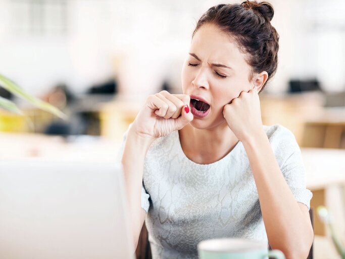 Junge Frau in Büro sitzt gähnend am Schreibtisch vor dem Laptop | © iStock / Neustockimages 