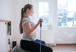 Schwangere Frau sitzt auf einem Gymnastikball mit Gummibändern in der Hand und trainiert Arme. | © Getty Images / Guido Mieth