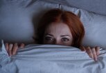 Rothaarige Frau liegt im Bett mit Bettdecke bis zur Nase gezogen und geöffneten Augen | © Getty Images/Axel Bueckert / EyeEm