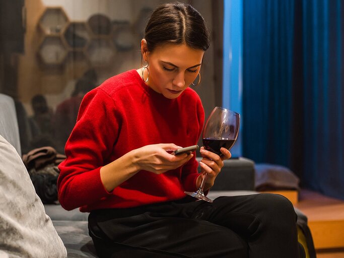 Frau mit einem Glas Rotwein in der Hand tippt auf ihrem Handy | © Getty Images/Rehulian Yevhen