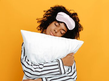  Afro-amerikanisches Mädchen mit Schlafbrille in Pyjama umarmt ein Kissen, auf gelbem Hintergrund. | © gettyimages.de | katiafonti