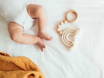 Baby liegt auf einem Bett, neben den Füßen ein Holzspielzeug und eine Decke. | © Getty Images / NelliSyr