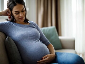 Hübsche Frau sitzt auf einem Sofa und streichelt ihren Babybauch | © Getty Images/damircudic