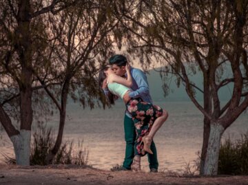 Ein Mann und eine Frau stehen an einem See unter Bäumen und küssen sich. | © Unsplash | Christian Newman