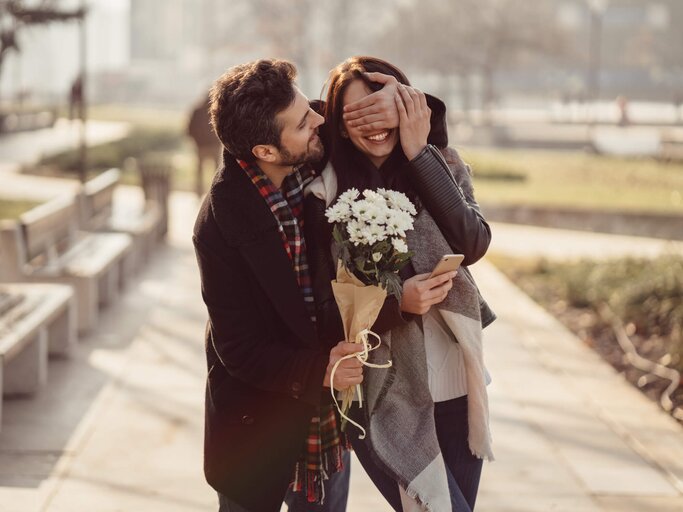 Mann führt seine Freundin mit geschlossenen Augen und Blumenstrauß in der Hand | © gettyimages.de | martin-dm