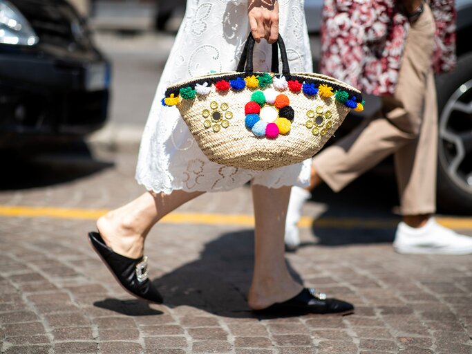  Streetstyle mit weißem Spitzenkleid, Basttasche mit bunten Bommeln und schwarzen Mules mit Straß-Detail auf der Mailänder Menswear Fashion Week | © Getty Images / Christian Vierig