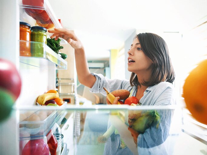 Brünette Frau hält frisches Obst und Gemüse im linken Arm und greift in die geöffnete Kühlschranktür. | © iStock | gilaxia