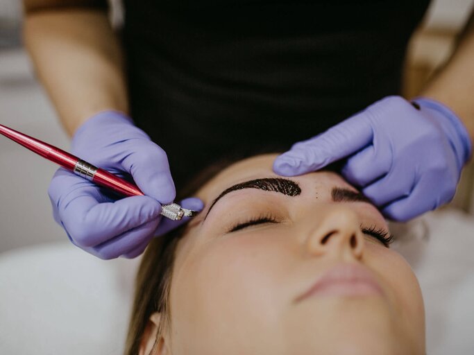 Frau liegt im Kosmetikstudio, während Kosmetikerin mit violetten Einmalhandschuhen mit Microblading-Skalpell ihre Augenbrauen verdichtet | © iStock | MMPhotography