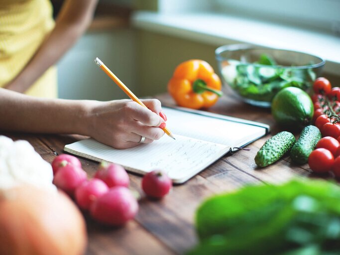 Frau notiert sich Ernährungsplan mit Bleistift in einem Notizbuch, das auf einer Holzarbeitsplatte zwischen verschiedenen Gemüsesorten liegt | © iStock | yulkapopkova