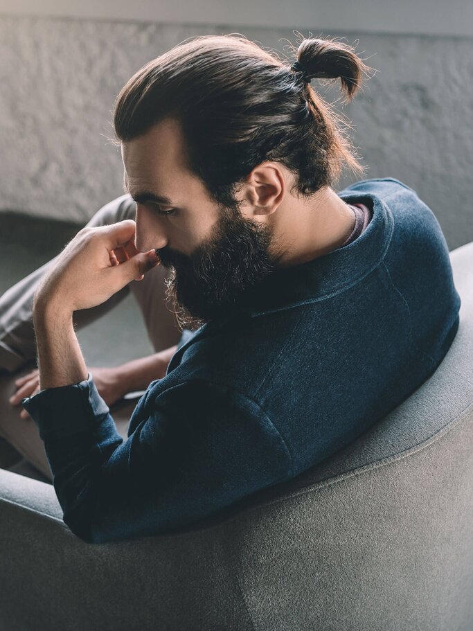 Ein Mann, der einen Man Bun und Vollbart trägt, sitzt auf einem Sessel. | © iStock | LightFieldStudios