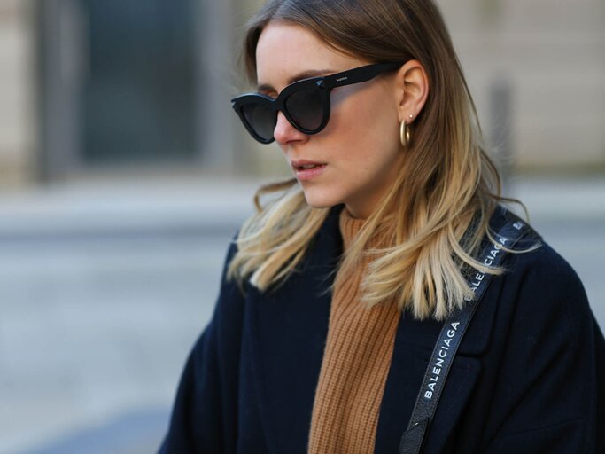 Junge Frau mit Sombre Hair in Aschblond mit hellblonden Spitzen trägt schwarze Sonnenbrille, einen beigen Pullover, einen schwarzen Mantel und eine Balenciaga Umhängetasche | © Getty Images | Jeremy Moeller 