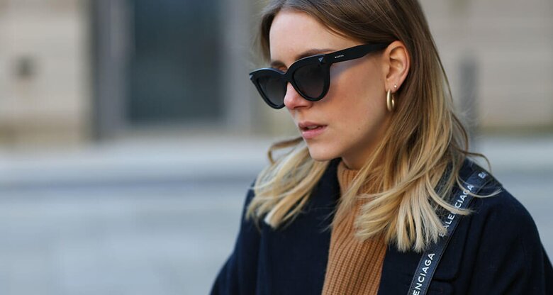 Junge Frau mit Sombre Hair in Aschblond mit hellblonden Spitzen trägt schwarze Sonnenbrille, einen beigen Pullover, einen schwarzen Mantel und eine Balenciaga Umhängetasche | © Getty Images | Jeremy Moeller 