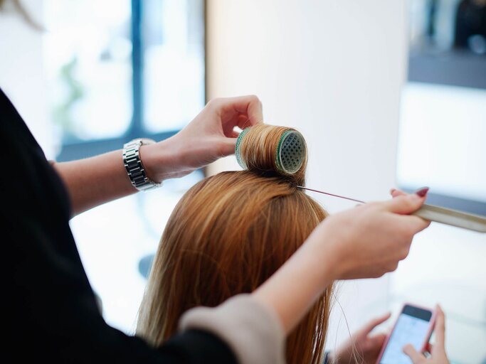 Junge Frau mit langen rötlichen Haaren bei Frisörin, die Lockenwickler in den Ansatz eindreht | © Getty Images | Caiaimage | Gianni Diliberto