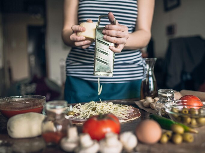 Frau mit Streifenshirt reibt Käse auf Pizza, die neben der Tomatensauce, Oliven, Eiern und Champignons auf Küchenarbeitsfläche liegt | © iStock | hobo_018