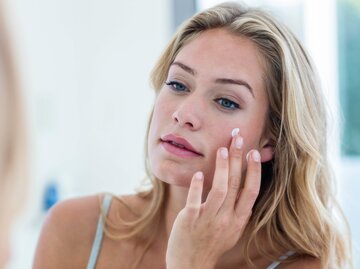 Junge Frau steht vor Spiegel und trägt Retinol Creme auf ihre Wange auf | © iStock | Wavebreakmedia