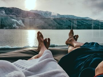 Frau und Mann liegen m Bademantel auf Liegestuhl am See mit Blick auf Bergpanorama | © iStock | Christopher Moswitzer