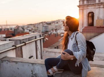 Junge Frau sitzt mit Rucksack und Sonnenbrille bei Sonnenuntergang auf Mauer einer Dachterrasse und genießt den Blick über Lissabon | © Getty Images | visualspace