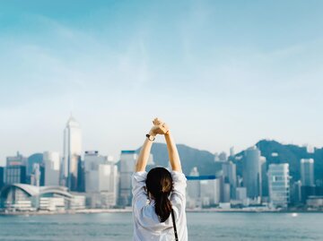 Brünette Frau mit Pferdeschwanz streckt beide Arme in die Luft und blickt auf Skyline von Hongkong | © Getty Images | d3sign