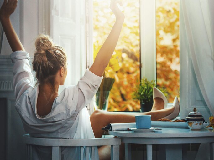 Frau sitzt am Küchentisch, Füße auf dem Tisch und streckt Arme nach oben | © iStock | gilaxia