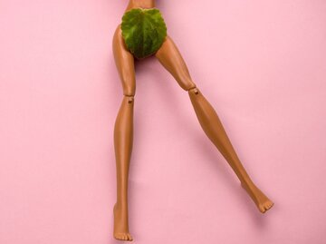 Beine einer Puppe auf Rosa Hintergrund, ein grünes Blatt bedeckt den Schambereich. | © iStock.com / dvulikaia