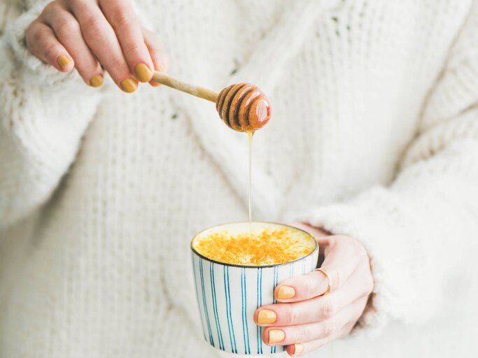 Junge Frau mit Wollpullover und gelben Nagellack träufelt Honig in eine Tasse mit Goldener Milch. | © iStock | Foxys_forest_manufacture