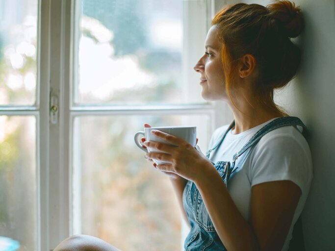 Junge schöne Frau schaut durch ein Fenster und trinkt Kaffee am Morgen | © iStock | Jasmina007
