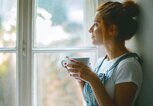 Junge schöne Frau schaut durch ein Fenster und trinkt Kaffee am Morgen | © iStock | Jasmina007