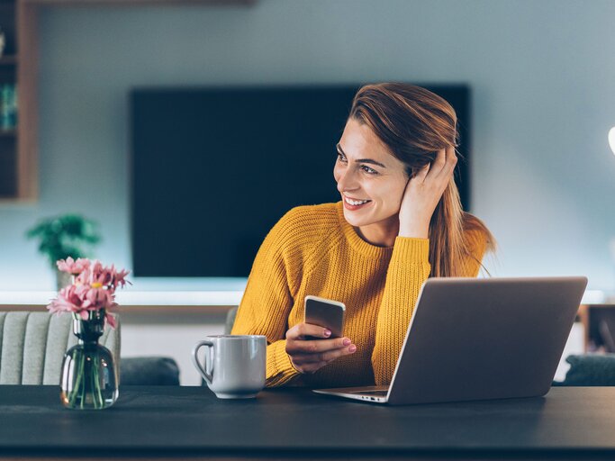 Junge Frau sitzt am PC mit ihrem Handy in der Hand | © iStock | filadendron