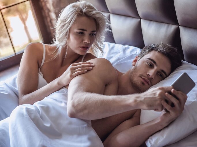 Junges Paar liegt im Bett, der Mann hält sein Handy in der Hand. | © iStock | GeorgeRudy