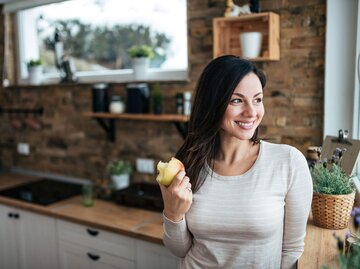 Gesunde Frau steht in der Küche am Fenster und isst einen Apfel. | © iStock | nortonrsx