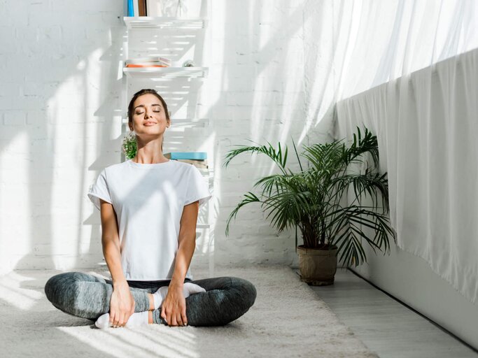 Junge Frau sitzt im Zimmer auf dem Boden im Schneidersitz und sieht gesund und glücklich aus. | © iStock | LightFieldStudios