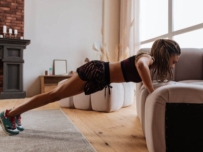 Frau in Sportkleidung macht Couch Push-Ups im Wohnzimmer am Sofa. | © iStock | yacobchuk