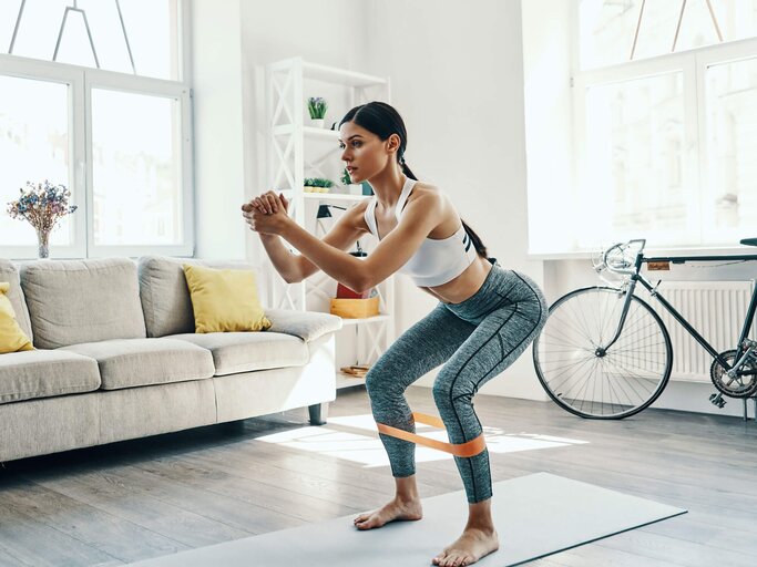 Junge Frau übt in Sportkleidung in ihrem Wohnzimmer Kniebeugen. | © iStock | g-stockstudio