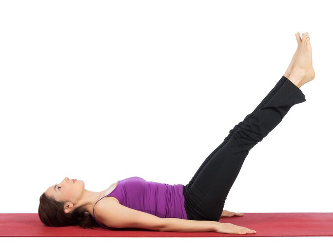 Frau liegt mit dem Rücken auf einer Yoga-Matte und hebt ihre Beine gerade an. | © iStock | DenizA