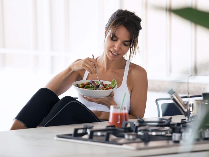 Junge Frau im Sportoutfit sitzt in der Küche und isst Salat. | © iStock | nensuria