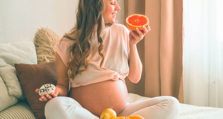 Schwangere Frau sitzt auf dem Bett mit Obst und Gemüse vor sich.  | © iStock.com / Anastasiia Stiahailo