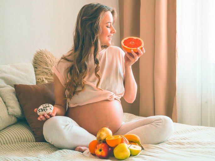 Schwangere Frau sitzt auf dem Bett mit Obst und Gemüse vor sich.  | © iStock.com / Anastasiia Stiahailo