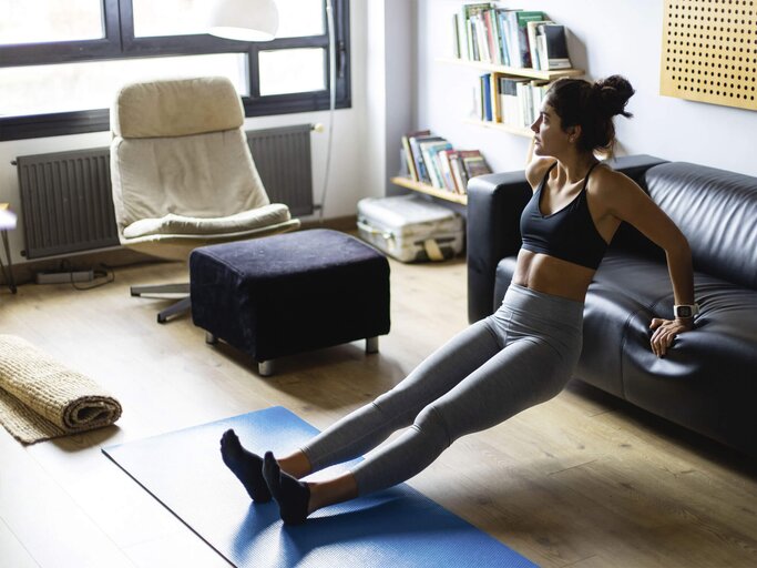 Junge Frau trainiert mit Trizeps-Dips zuhause im Wohnzimmer ihre Oberarme. | © iStock.com / Nando Martinez