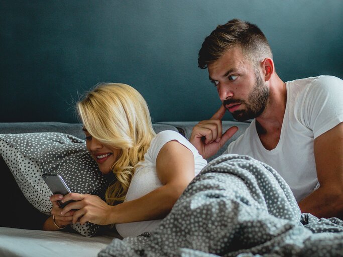 Junges Paar liegt im Bett, sie schaut auf ihr Handy | © iStock.com / blackCAT