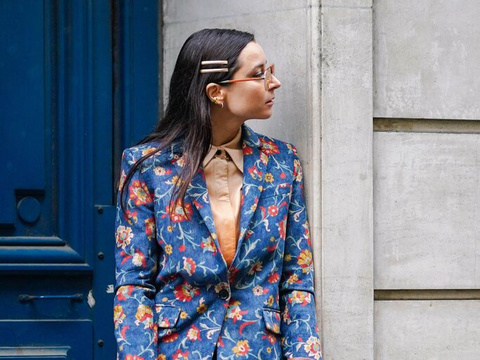 Frau steht in einem Hauseingang, trägt einen blauen Blazer mit Blumenprint und hat Haarspangen in den Haaren. | © gettyimages.de / Edward Berthelot