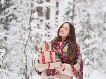 Frau steht im Schnee mit Weihnachtsgeschenken auf dem Arm.  | © iStock.com / Maya23K