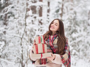 Frau steht im Schnee mit Weihnachtsgeschenken auf dem Arm.  | © iStock.com / Maya23K