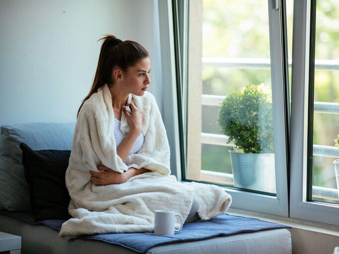 Frau sitzt mit Halsschmerzen auf der Couch und schaut aus dem Fenster. | © gettyimages.de / milanvirijevic