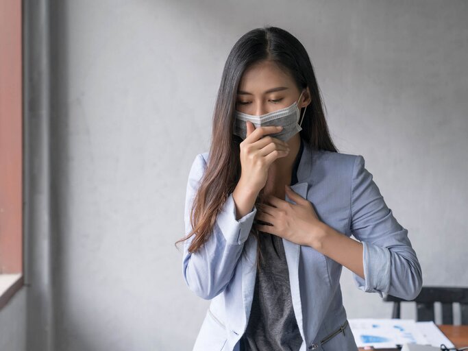 Frau mit Mundschutz hält die Hand vor den Mund und hustet. | © gettyimages.de / thianchai sitthikongsak