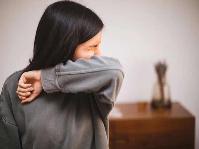 Frau mit einem grauen Pullover niest sich in die Ellenbeuge.  | © gettyimages.de / agrobacter