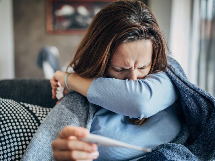 Frau sitzt warm eingepackt auf der Couch und hält ein Fieberthermometer in der Hand. | © gettyimages.de / South_agency