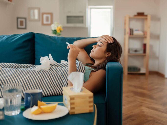 Frau liegt mit einer Erkältung auf der Couch, fühlt mit der Hand die Stirn und hält ein Fieberthermometer.  | © gettyimages.de / PixelsEffect