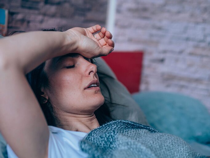 Frau liegt mit Fieber und geschlossenen Augen im Bett und hält ihre Stirn. | © gettyimages.de / VioletaStoimenova