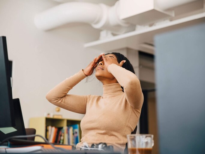 Frau sitzt am Computer, lehnt sich im Stuhl zurück und hält sich aufgrund von Kopfschmerzen den Kopf. | © gettyimages.de / Maskot