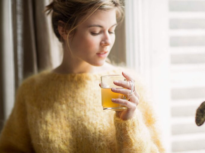 Frau mit gelben Pullover steht am Fenster und trinkt eine Tasse Tee. | © gettyimages.de / Letizia Le Fur